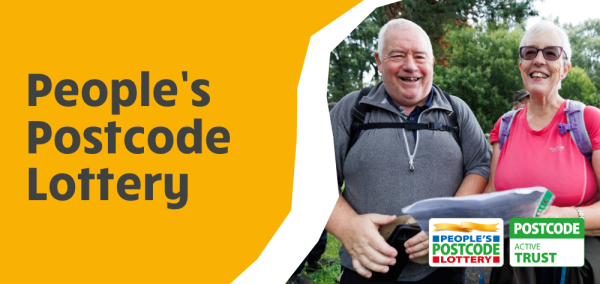 Two older adults with backpacks smile while looking at a map during a group walk in a green outdoor setting, representing the walking activities supported by People’s Postcode Lottery funding.